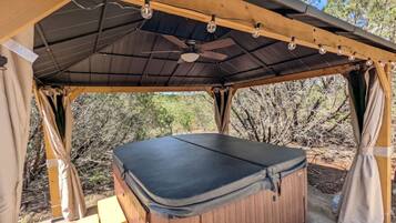 Een hottub onder een houten pergola, met een plafondventilator, omringd door bomen.
