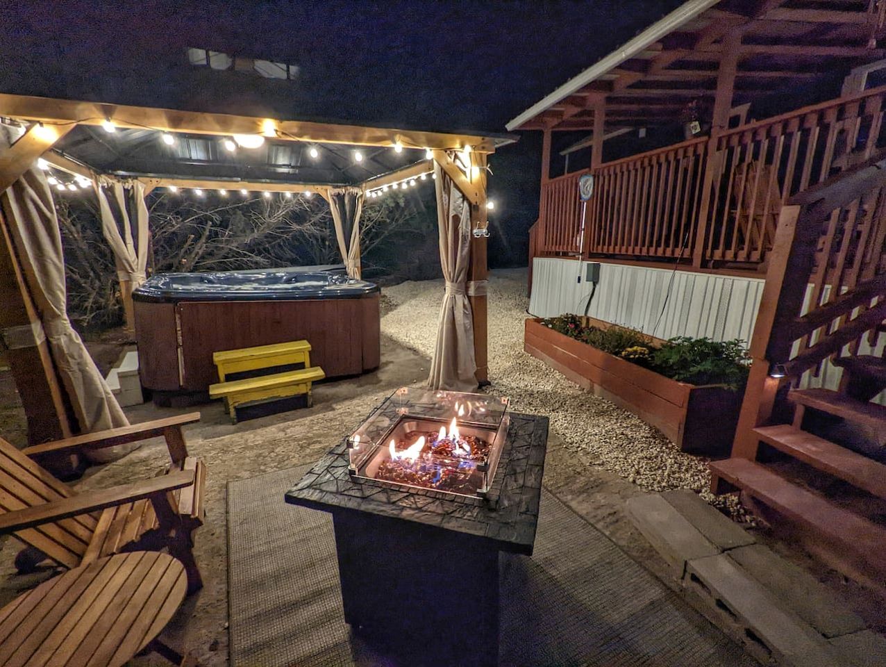 Un patio al aire libre con un pozo de fuego, asientos de madera, un jacuzzi y luces colgantes.