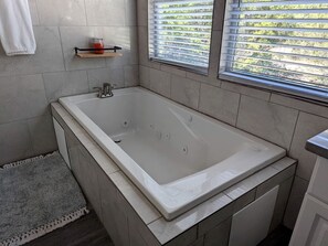 A modern bathroom with a large bathtub, tiled walls, a small shelf with a candle, and a window with blinds. - Romantic Stays in Texas at this Granbury Cabin Glamping Site (Granbury)