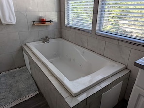 Une salle de bain moderne avec une grande baignoire, des murs carrelés, une petite étagère avec une bougie et une fenêtre avec des stores.