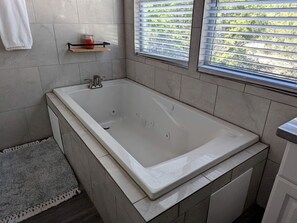 Et moderne badeværelse med et stort badekar, flisebeklædte vægge, en lille hylde med et lys og et vindue med persienner.