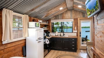 A compact kitchen with a refrigerator, microwave, and stove, wooden walls, a window with blinds, and a view of a lake.