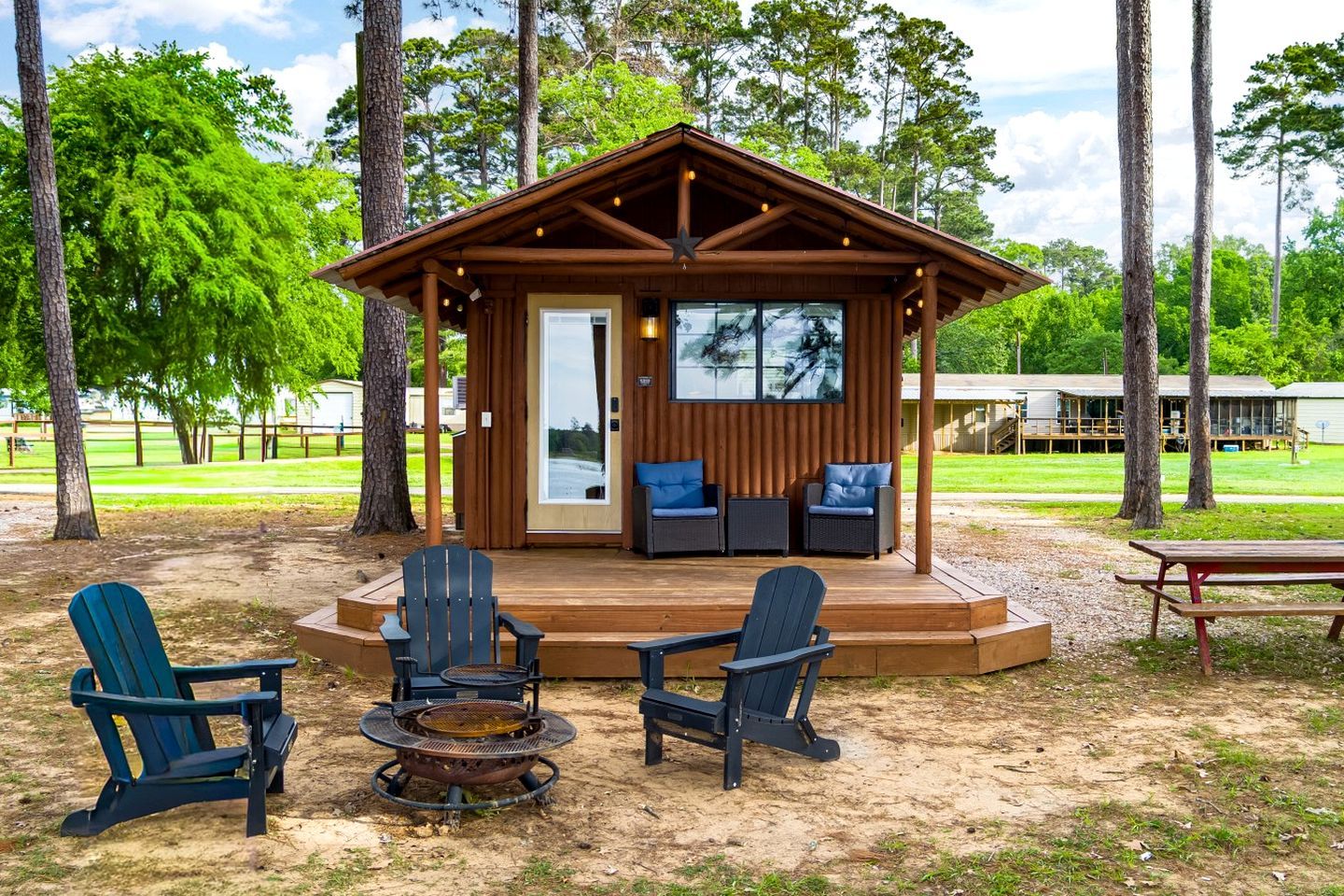 A wooden cabin with a deck, four Adirondack chairs, a fire pit, and a covered seating area with two sofas.