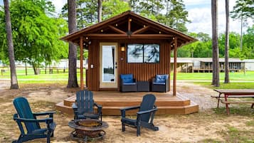 A wooden cabin with a deck, four Adirondack chairs, a fire pit, and a covered seating area with two sofas.