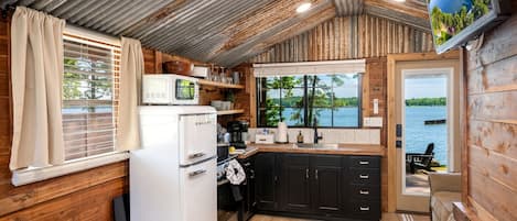 Cocina compacta con refrigerador, microondas y estufa, paredes de madera, ventana con persianas y vista a un lago.