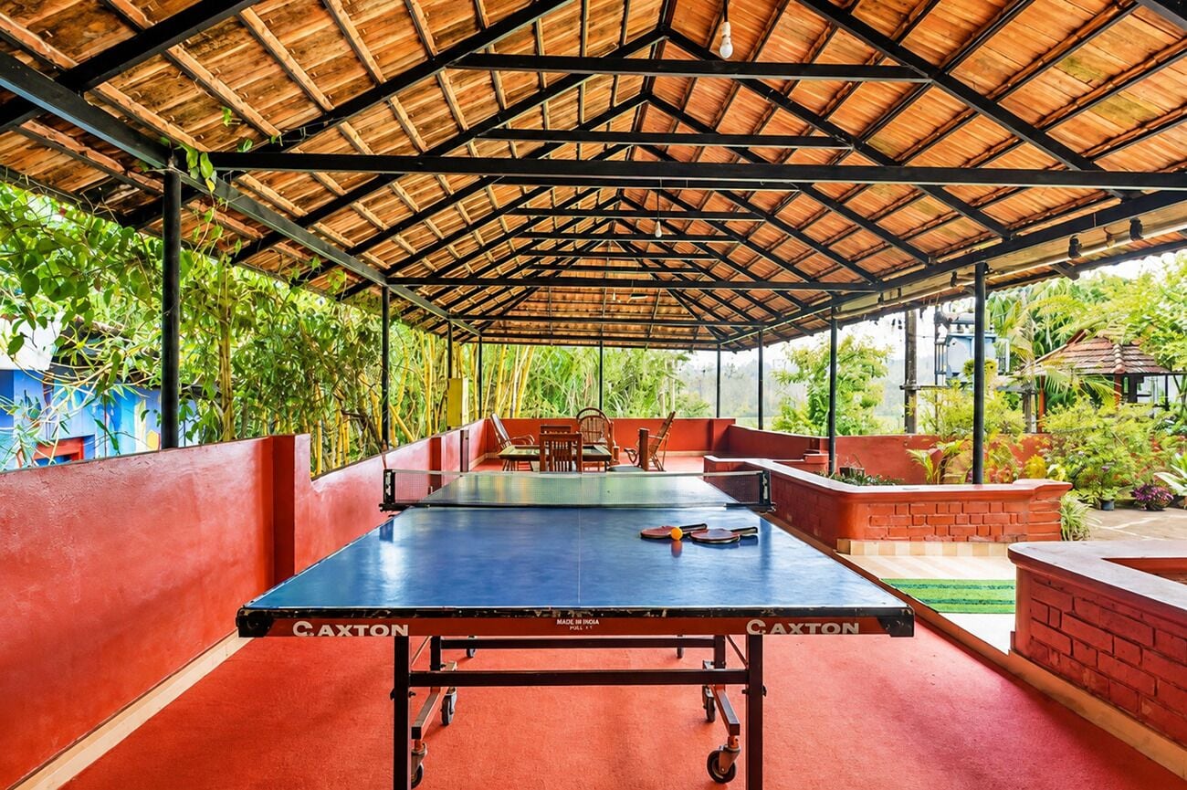 Una mesa de ping pong en el interior, con piso rojo, rodeada de asientos y un espacio cubierto con techo de madera.