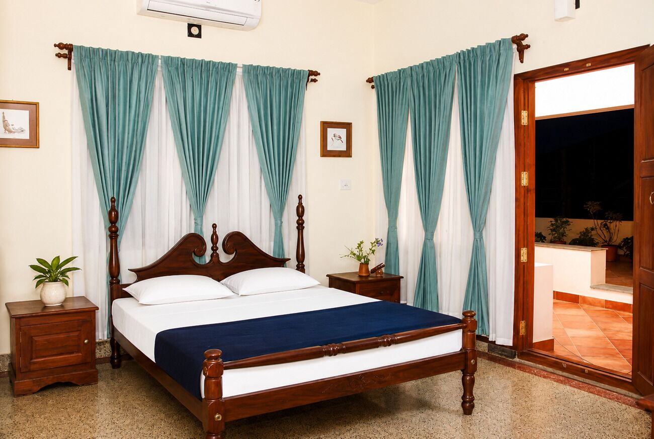 Un dormitorio con cama de madera, cortinas azul verdosas, una mesita de noche con una planta y vistas a un espacio empedrado.