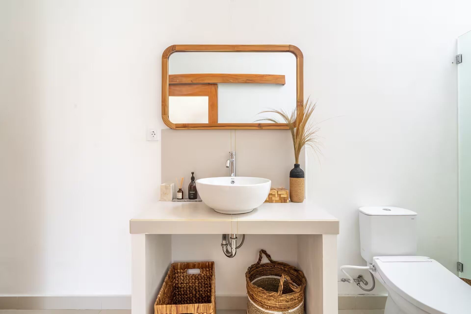 Un baño con lavabo blanco, espejo e inodoro. Hay cestas de mimbre debajo del lavabo y un florero con plantas secas en la encimera.