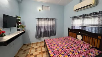 A bedroom with a bed, a mounted TV, an air conditioner, a small shelf with plants, and a patterned floor.