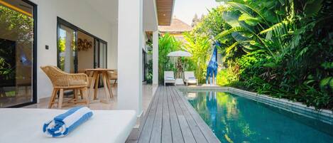 A modern pool area with a wooden deck, lounge chairs, and lush greenery.