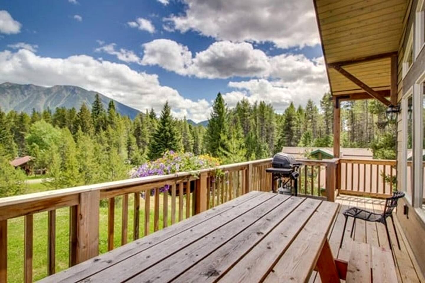 A wooden deck with a grill and a table, overlooking a forest and mountains.