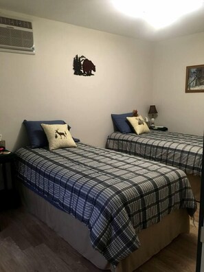 Two twin beds with checkered bedding, a wall-mounted heater, a small nightstand with a lamp, and a framed picture on the wall.