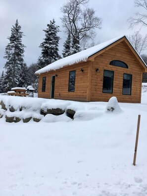 Una cabaña de madera con techo nevado, rodeada de árboles cubiertos de nieve y un muro de piedra.