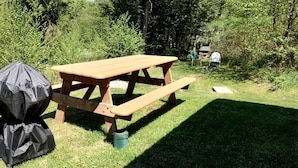 Ein Picknicktisch mit Grill und einem abgedeckten Gegenstand, umgeben von viel Grün.