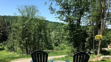 Zwei Adirondack-Stühle und ein kleiner Tisch in einer grünen Fläche mit einem Feldweg und Bäumen im Hintergrund.