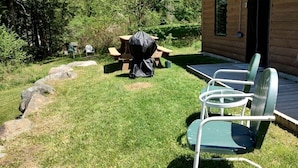 Eine Holzhütte mit einem überdachten Picknicktisch und Grill, einer überdachten Terrasse mit Stühlen sowie einer Rasenfläche mit Steinen.