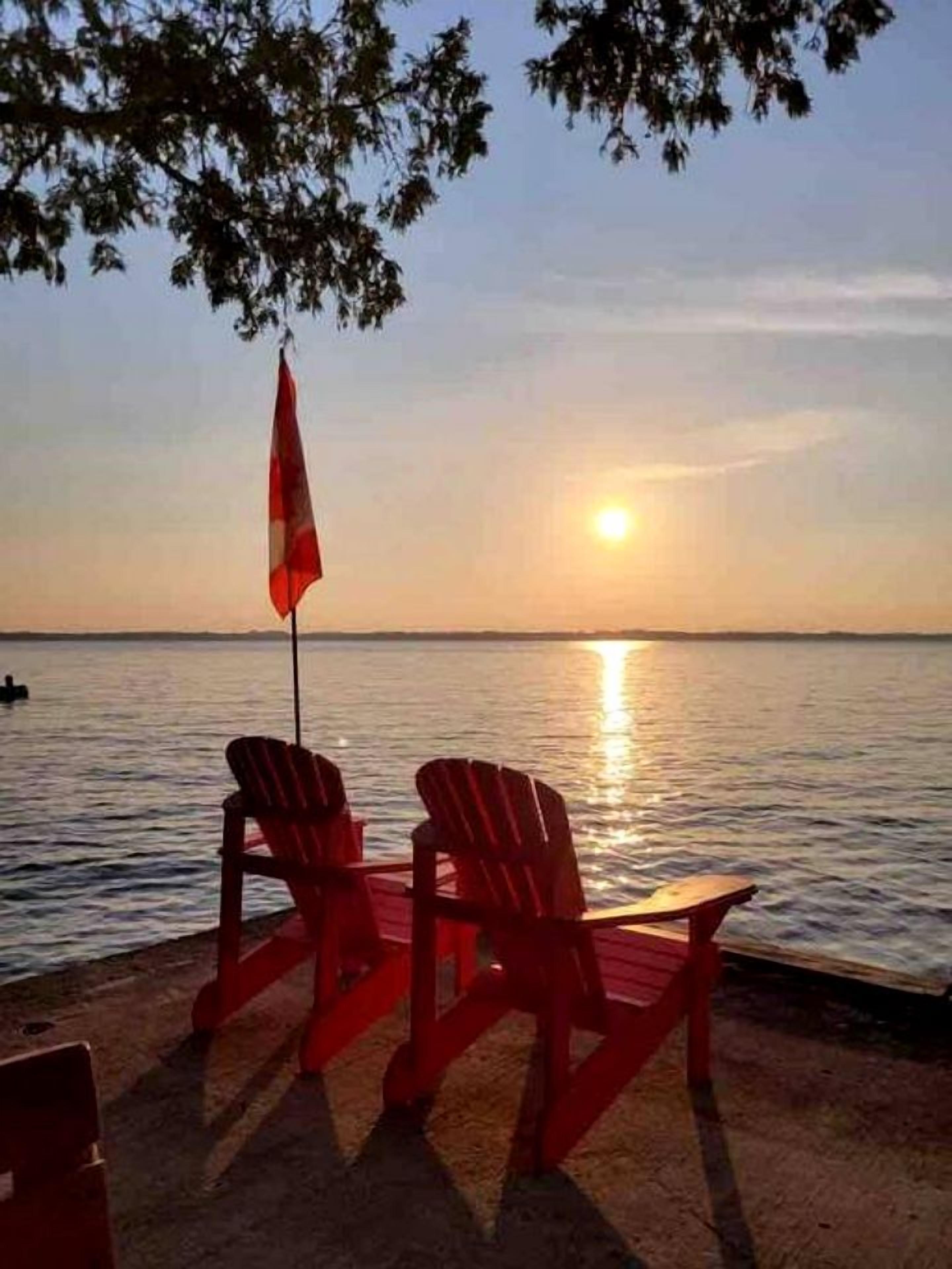 Two Adirondack chairs facing a sunset over a body of water.