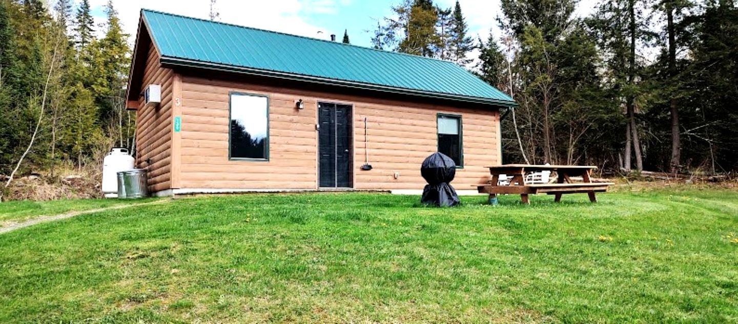 A log cabin with a green roof, a picnic table, and a trash can in front.