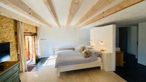 A bedroom with a wooden ceiling, a bed with pillows, a nightstand with lamps, a TV, and a stone wall. - La Grange du Sotoul - Large house for 10 people with all comforts and private swimming pool (Soturac)