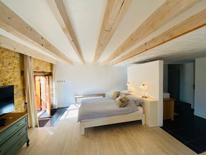 Une chambre avec un plafond en bois, un lit avec des oreillers, une table de chevet avec des lampes, une télévision et un mur en pierre.