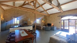 A rustic interior with exposed wooden beams, a kitchen with a central island, a dining area with a table and chairs, a living area with sofas, and a large window providing natural light. - La Grange du Sotoul - Large house for 10 people with all comforts and private swimming pool (Soturac)