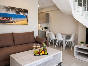 Ein modernes Wohnzimmer mit einem braunen Sofa, einem Essbereich mit weißen Stühlen, einer Küche im Hintergrund und einem Gemälde mit einer Strandansicht.