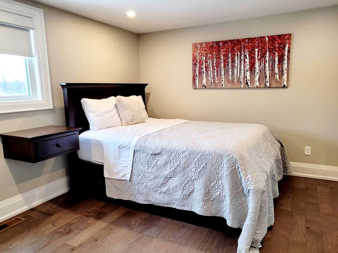 A bedroom with a bed, a nightstand, a wall-mounted painting, and a window.