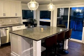 Una cocina con isla central, taburetes de bar, iluminación colgante y vistas al exterior.