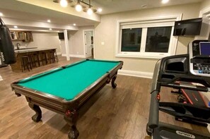Una mesa de billar en una habitación con una cinta de correr, una zona de bar y una ventana con persianas.