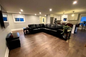 A living room with a black leather sectional sofa, a small side table, a potted plant, and a kitchen in the background. - Spacious Lakefront Cottage with Private Boat Launch on Chemong Lake, Near Peterborough, Ontario (Harcourt)