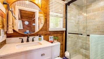 A bathroom with a wooden wall, a round mirror, a sink with two faucets, a toilet, and a glass shower enclosure.