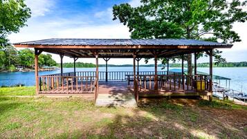 A wooden gazebo with a metal roof overlooking a lake, surrounded by trees and a dock.