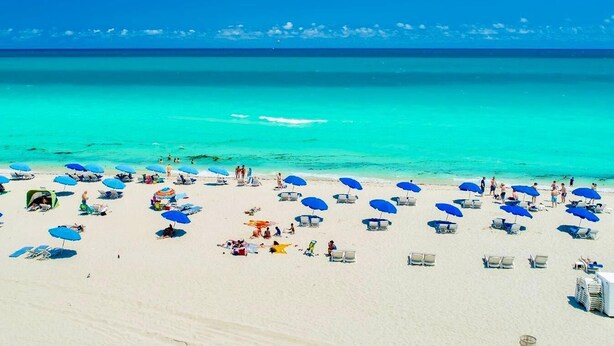 Una playa con numerosos sombrillas y tumbonas, un cielo azul despejado y el océano.
