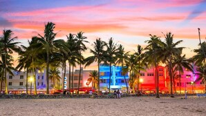 Un hotel frente a la playa, con palmeras, un muro de piedra y un cielo al atardecer lleno de colores.