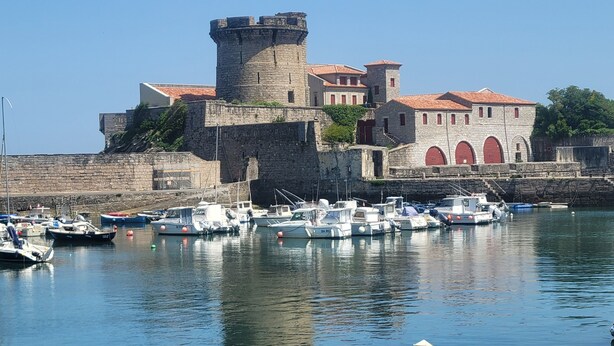 Un bâtiment historique en pierre, doté d’une tour et surmonté d’un toit rouge, ainsi qu’un port de plaisance avec des bateaux.