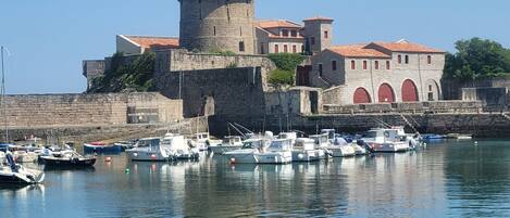 Un edificio de piedra histórico con una torre, una estructura de techo rojo y un puerto deportivo con barcos.