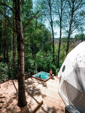 A forested area with a wooden deck, a hot tub, and a covered area.