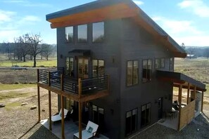 A modern two-story house with a balcony, large windows, and a covered outdoor seating area. - Modern Luxury Perfect for Family Getaways with Game Room, Hot Tub & Fire Pit in Broken Bow, Oklahoma (Broken Bow)