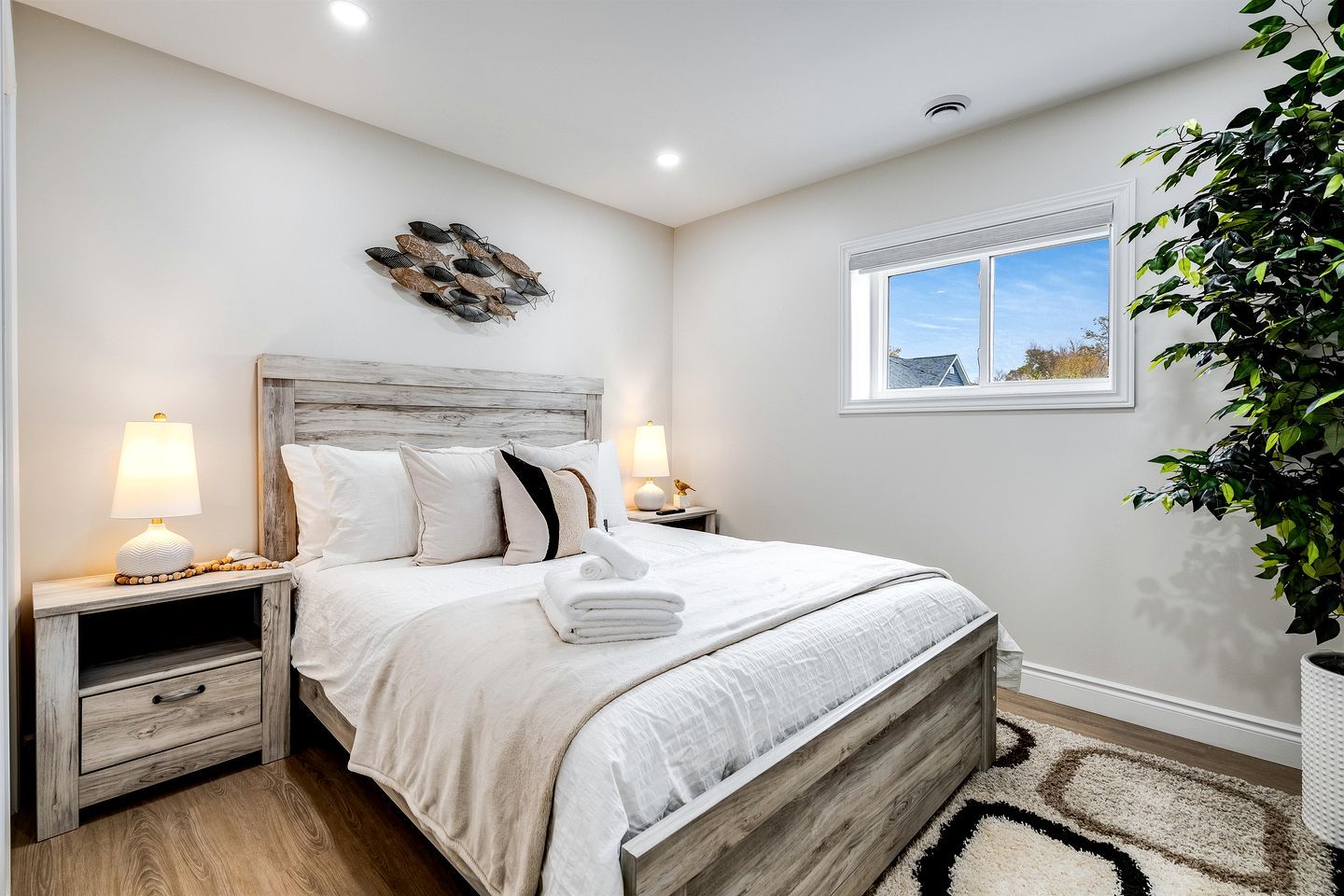 A bedroom with a wooden headboard, a bed with white linens, two bedside lamps, a nightstand with a drawer, a window with a view, and a potted plant.