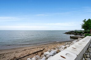 A beach with a concrete barrier, calm water, and a distant rocky outcrop. - Luxurious Beachfront Rental with Hot-Tub and Yard in Canada (Kingsville)