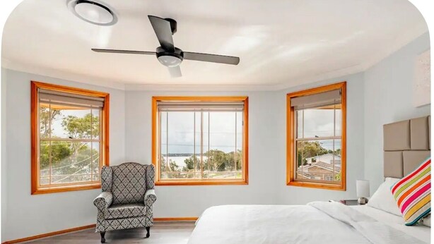 Ein Schlafzimmer mit Bett, Stuhl, Deckenventilator und drei Fenstern mit Blick.