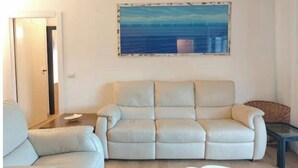 A living room with a beige sofa, a white armchair, a glass coffee table, and a painting of a blue sea on the wall. - Hostdomus - Myros Apartment (Finale Ligure)
