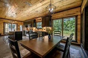 A wooden cabin interior with a dining area, kitchen, and large windows. - Surrounded by Tall Pines Forest Views with Hot Tub, Game Room & Outdoor Fireplace in Broken Bow, Oklahoma (Broken Bow)