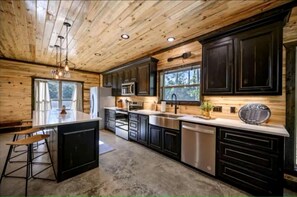 A modern kitchen with wooden ceiling, dark cabinets, stainless steel appliances, and a bar area. - Surrounded by Tall Pines Forest Views with Hot Tub, Game Room & Outdoor Fireplace in Broken Bow, Oklahoma (Broken Bow)