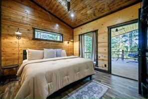 A wooden cabin bedroom with a bed, nightstands, a window, and a view of the outdoors. - Surrounded by Tall Pines Forest Views with Hot Tub, Game Room & Outdoor Fireplace in Broken Bow, Oklahoma (Broken Bow)