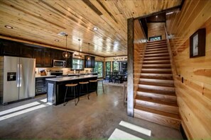 A modern kitchen with a central island, stainless steel appliances, and a wooden staircase. - Surrounded by Tall Pines Forest Views with Hot Tub, Game Room & Outdoor Fireplace in Broken Bow, Oklahoma (Broken Bow)