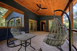 A wooden deck with hanging chairs and a table, a ceiling fan, and a view of a house with large windows. - Surrounded by Tall Pines Forest Views with Hot Tub, Game Room & Outdoor Fireplace in Broken Bow, Oklahoma (Broken Bow)