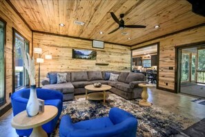 A log cabin interior with a large sofa, two blue armchairs, a coffee table, a ceiling fan, and a dining area visible through glass doors. - Surrounded by Tall Pines Forest Views with Hot Tub, Game Room & Outdoor Fireplace in Broken Bow, Oklahoma (Broken Bow)