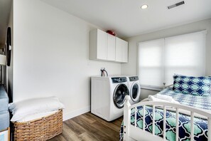 Een wasruimte met een wasmachine en droger, een bed met decoratieve beddengoed, een rieten mand en een wit kastje.