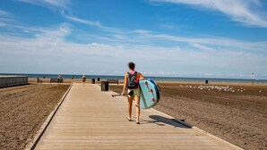 En träpromenad som leder till en strand, och på den går en person med en surfbräda.